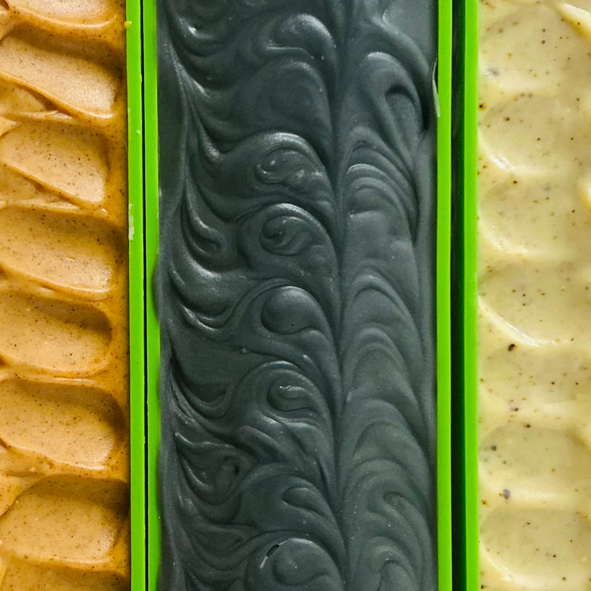 Three rectangular bars of soap in bright green molds, shown in a close-up vertical layout. The center bar is dark charcoal gray with a marbled swirl pattern, flanked by two tan and cream speckled bars.