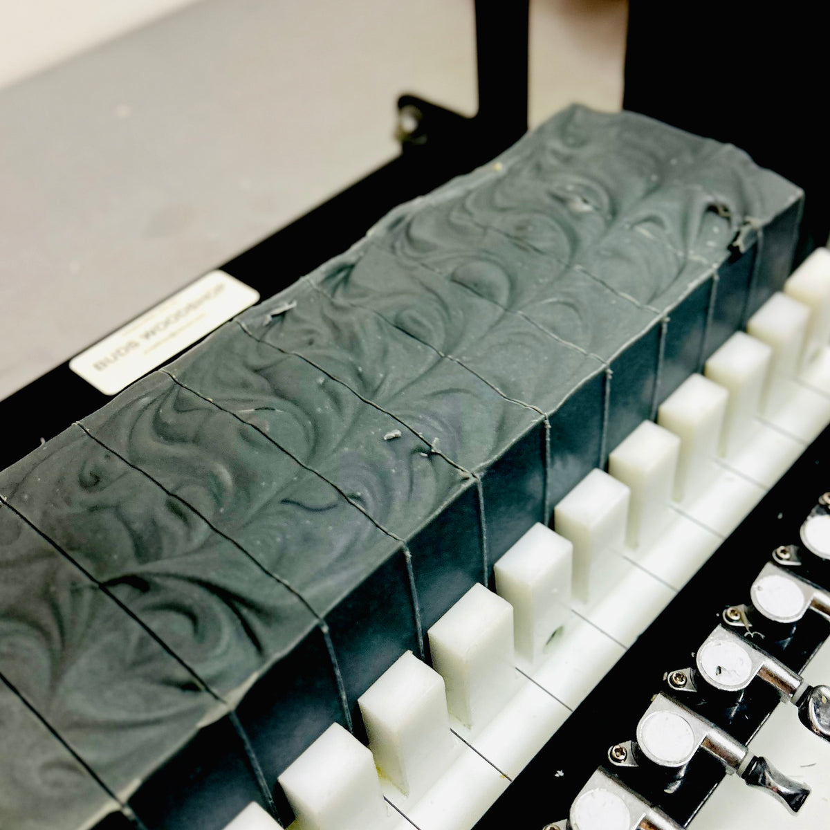 A close-up of a long rectangular bar of soap with a dark gray and black swirled pattern, shown resting on a white and black scale or platform with white blocks.