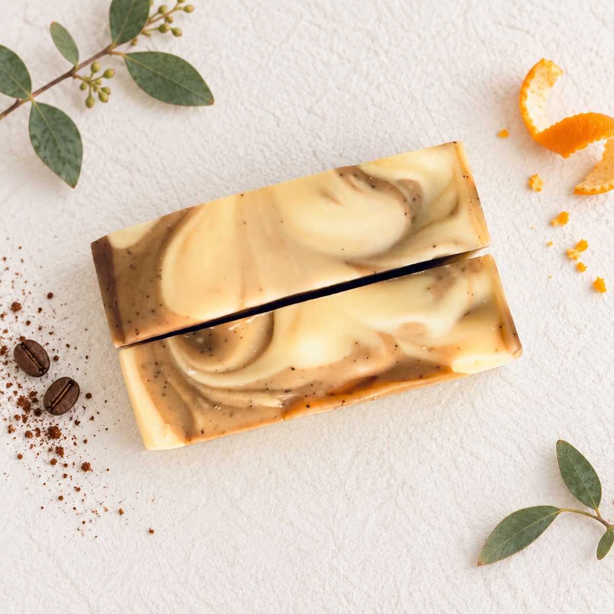 Two rectangular artisan soap bars with a marbled cream, brown, and beige swirl pattern, styled on a textured white background with coffee beans, cocoa powder, eucalyptus leaves, and an orange peel.