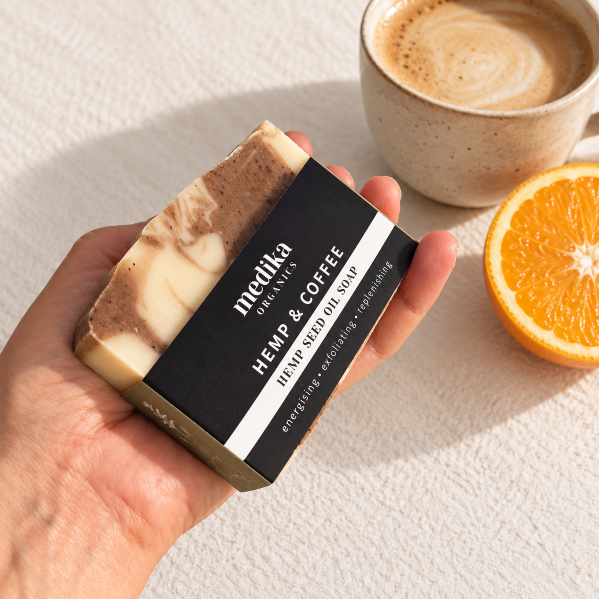 A hand holds a bar of Medika Organics Hemp & Coffee Hemp Seed Oil Soap, styled with a cup of coffee and a slice of orange on a textured white surface.