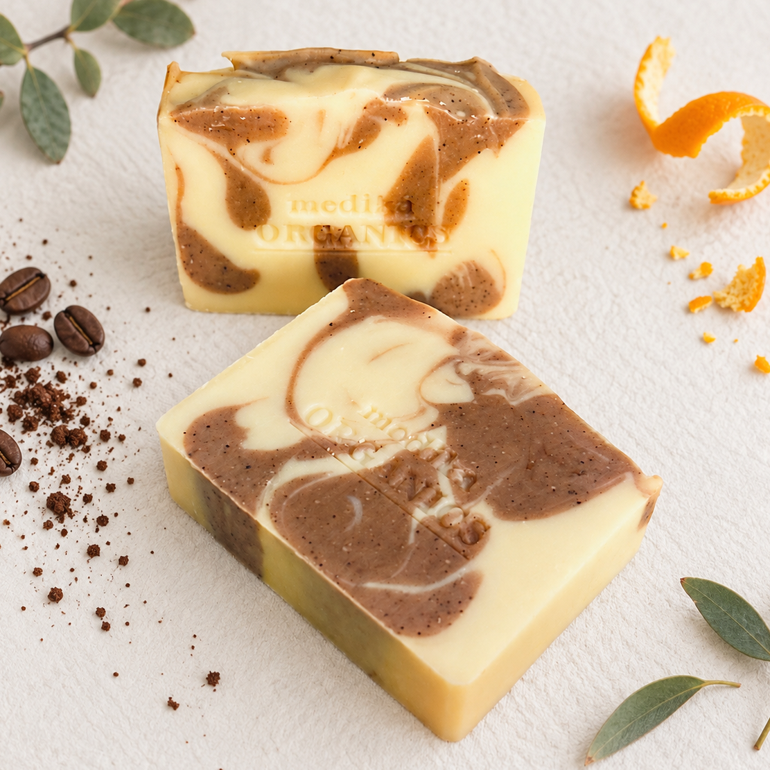 Two artisanal soap bars with a cream and brown marbled swirl pattern, styled with coffee beans, cocoa powder, orange peel, and green leaves on a textured white background. The brand name 'MEDIKA ORGANICS' is embossed on the soaps.