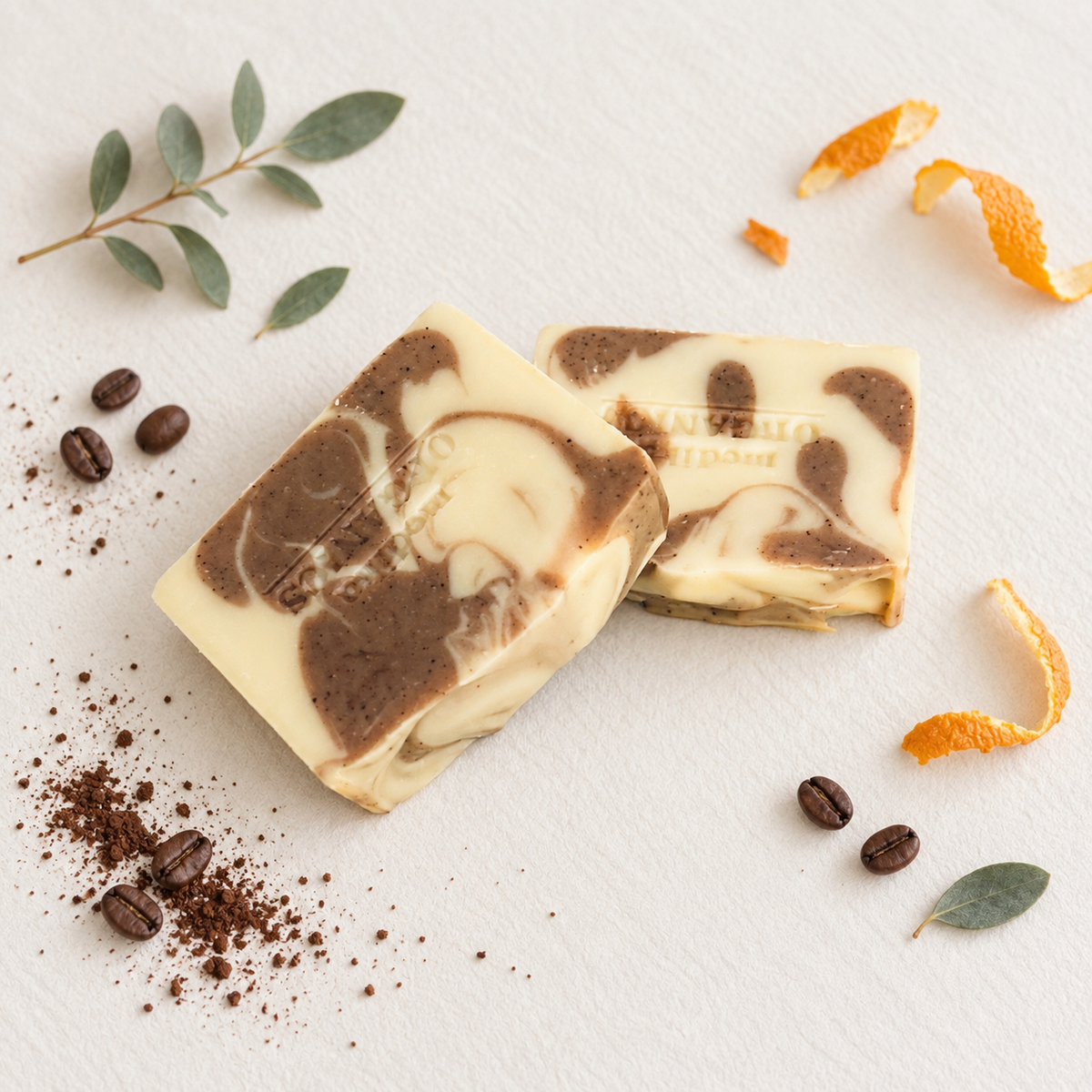 Two rectangular bars of marbled soap in cream and dark brown, styled with coffee beans, coffee powder, orange peel, and green leaves on a textured white background.