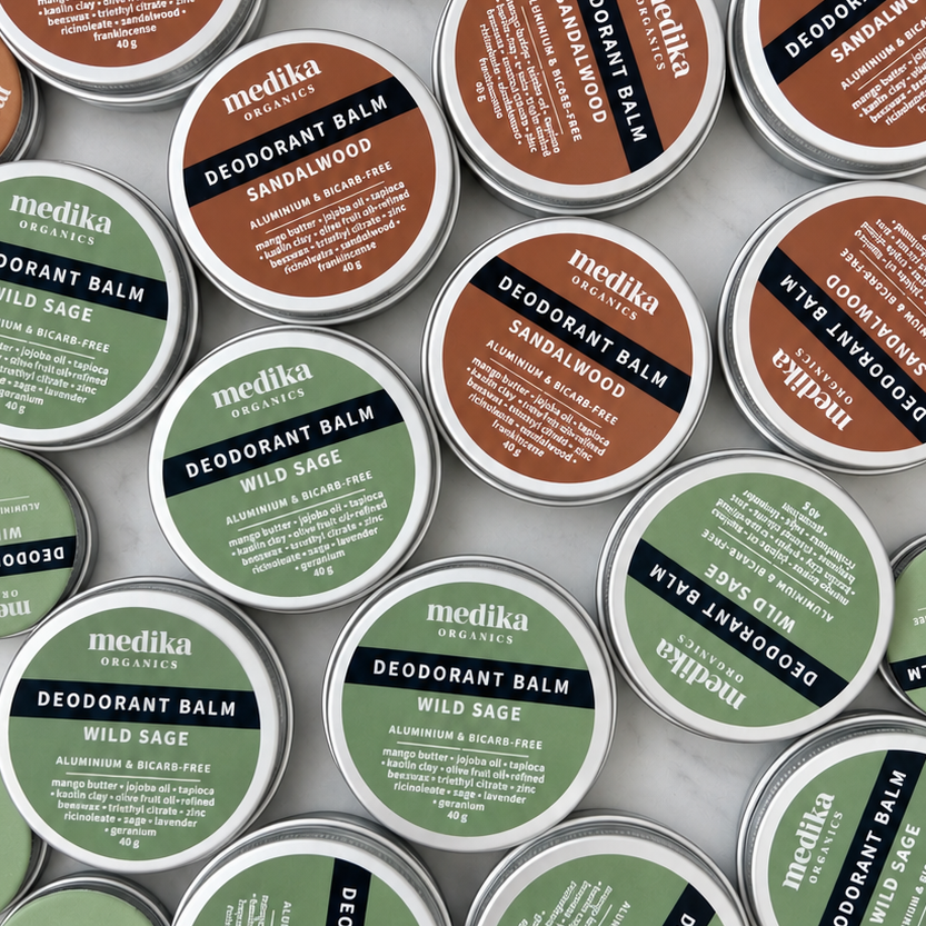 Medika Organics Deodorant Balms arranged in a flat lay on a light surface. The tins have silver rims, with round brown, sage green, and olive green lids featuring white text.