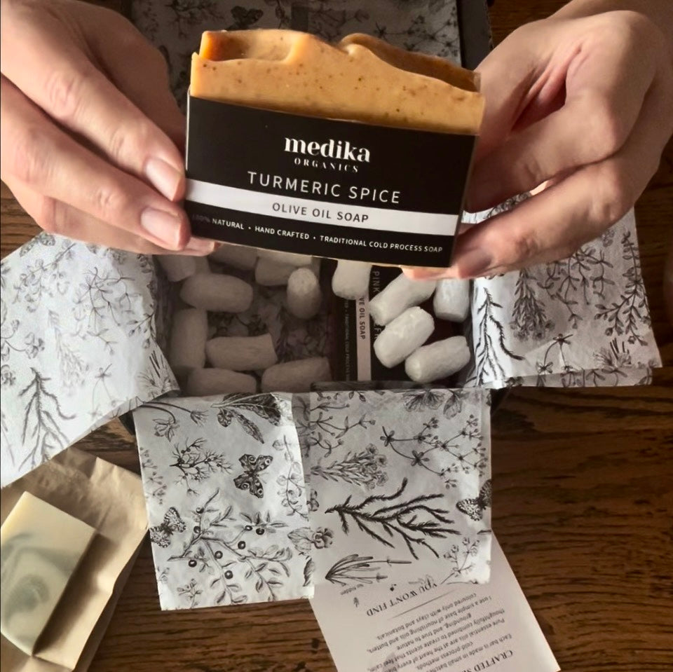 Hands holding a bar of Medika Organics Handmade Turmeric Spice Olive Oil Soap above an open gift box filled with botanical-patterned wrapping paper on a wooden table.