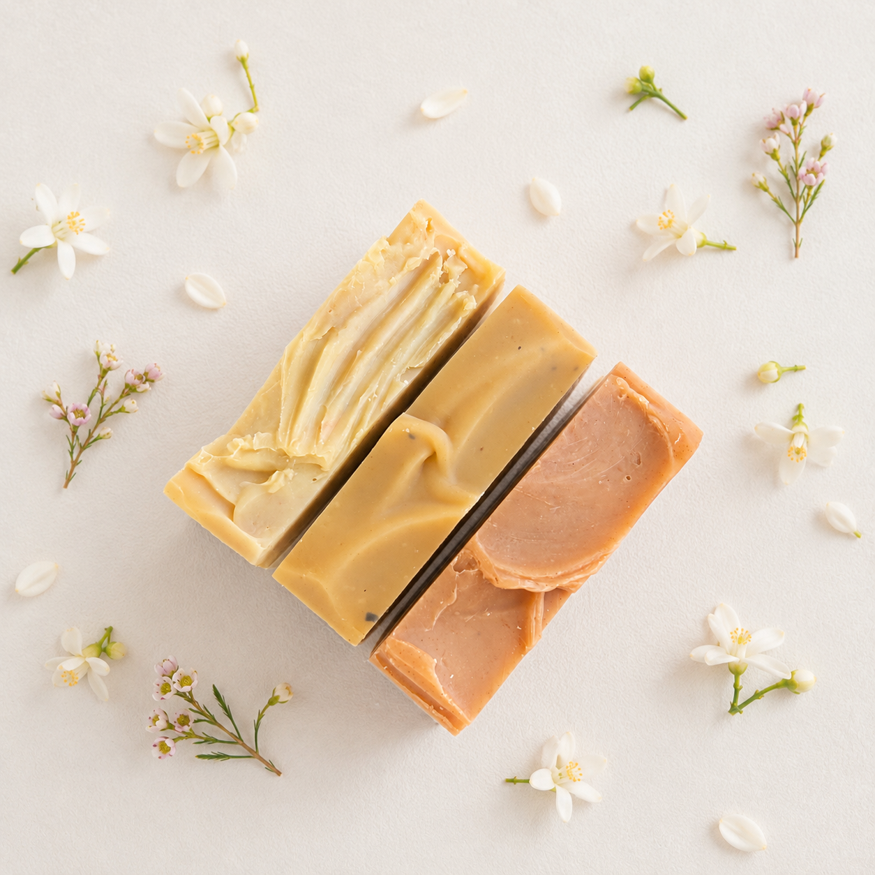 Three rectangular bars of handmade soap in pale yellow, beige, and peach tones, arranged diagonally on a light cream background with scattered white blossoms and small green stems.
