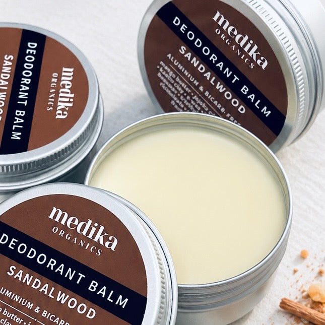 Three silver metal tins of Medika Organics Deodorant Balm in Sandalwood scent, with two closed tins showing brown labels and one open tin displaying the pale cream balm inside, set on a light surface.