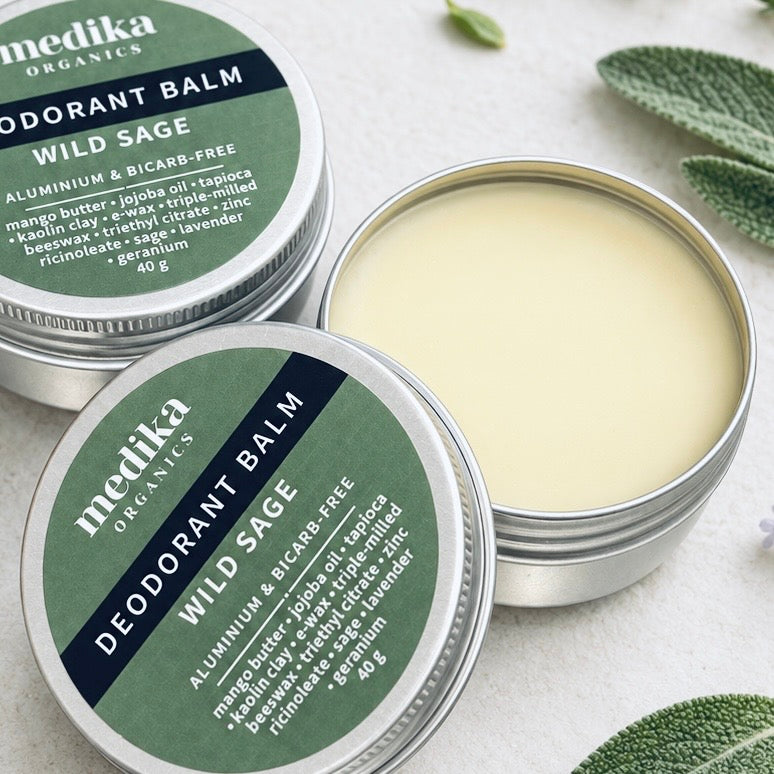 Medika Organics Wild Sage Deodorant Balm on a light textured surface, with one tin open to show the pale yellow balm inside and the other closed showing the green label.