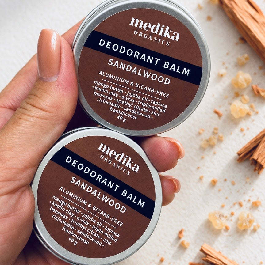 A hand holds two round metal tins of Medika Organics Deodorant Balm in Sandalwood scent.