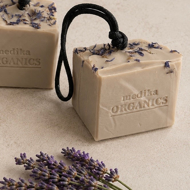 Soap On A Rope Bars of Medika Organics soap with dried lavender buds on top and black hanging loops, shown on a light stone surface with lavender sprigs in the foreground.