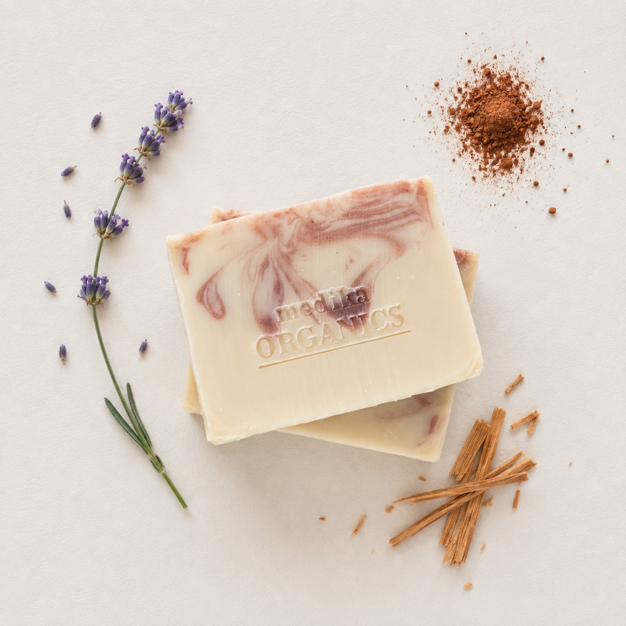 Two stacked bars of Medika Organics lavender and cinnamon soap on a light beige background, styled with a sprig of lavender, cedarwood and cocoa powder.