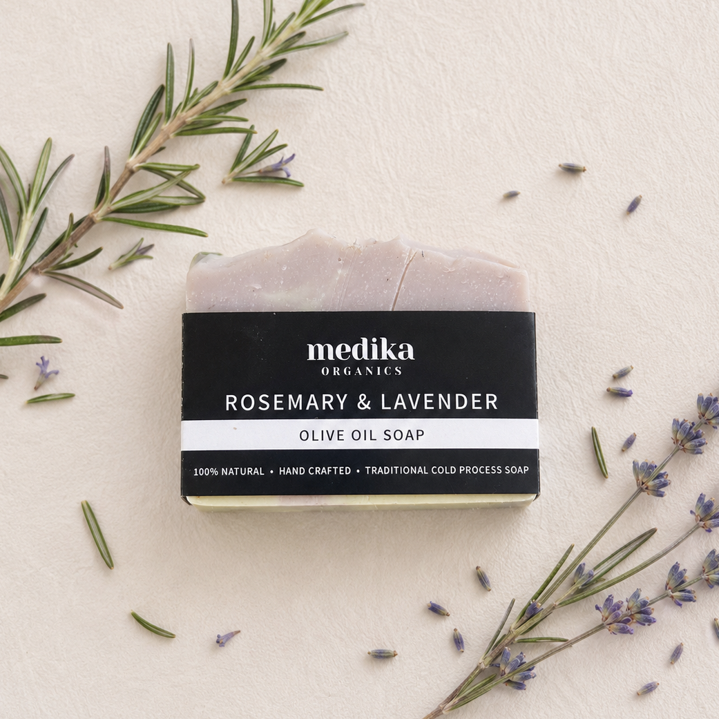 A bar of Medika Organics Rosemary & Lavender Olive Oil Soap is displayed on a textured cream background, styled with fresh rosemary sprigs and lavender buds. The soap bar has a light grayish-purple hue and is wrapped in a black label with white text.