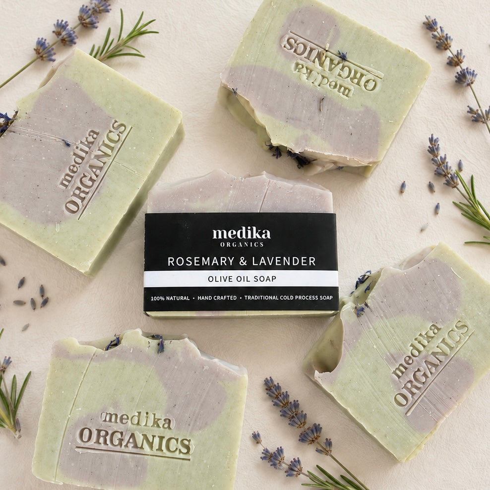 Five bars of Medika Organics Rosemary & Lavender Olive Oil Soap are arranged in a flat lay on a light beige surface, surrounded by sprigs of rosemary and lavender. One bar in the center has a black paper label wrapper, while the others show the natural marbled soap with a light green and lavender-gray color mix.