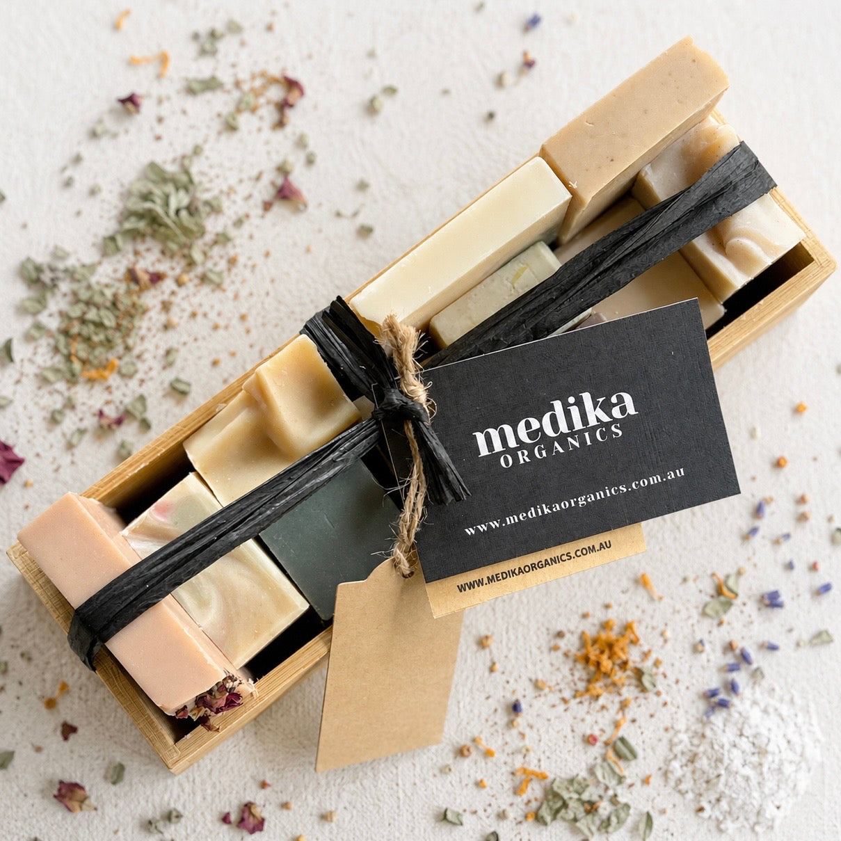 A set of handmade organic soap bars in a light wood box, wrapped with a black ribbon and a black paper tag reading 'medika organics'. The soaps are in shades of cream, beige, and light green, with dried botanicals scattered around on a white surface.