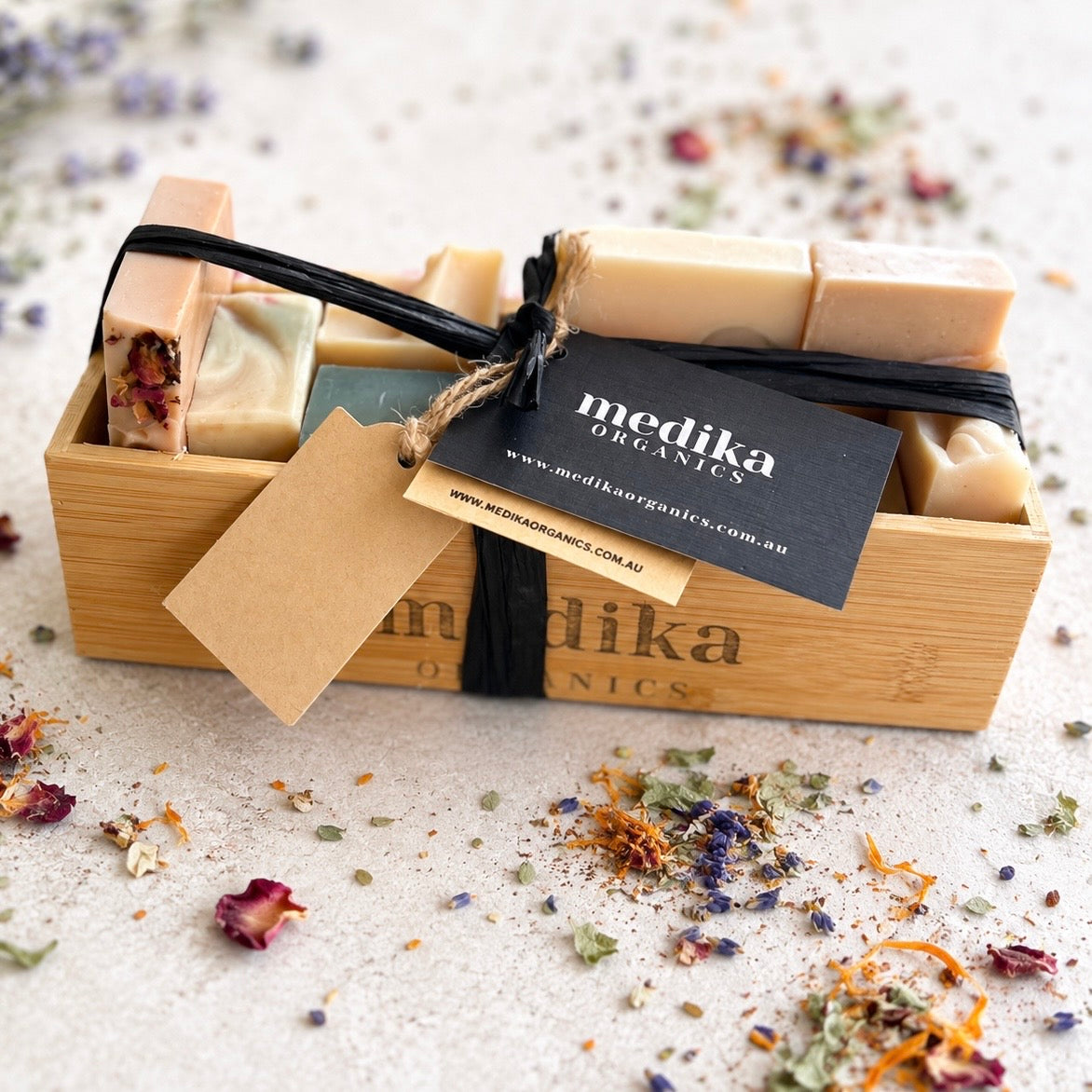 A wooden gift box containing several handmade soap bars tied with a black ribbon and twine, with a black MediKa Organics tag hanging from the front. The soaps are arranged inside a bamboo-style box on a light textured surface with dried flower petals scattered around.