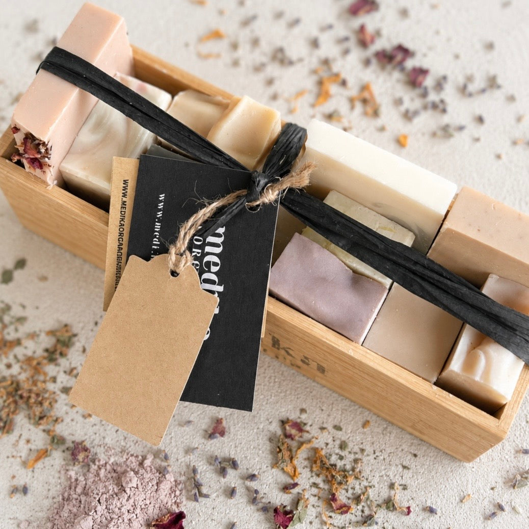 A wooden gift box containing several rectangular bars of soap in soft cream, beige, and pale lavender tones, tied with a black ribbon and kraft paper tags. Dried flower petals are scattered around on a light fabric background.