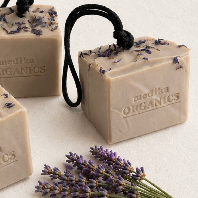Soap On A Rope bar of Medika Organics lavender soap in a pale lavender-gray color, decorated with dried lavender buds and a small white flower, complete with a black hanging loop. The soap is styled on a neutral beige textured surface with scattered lavender sprigs.