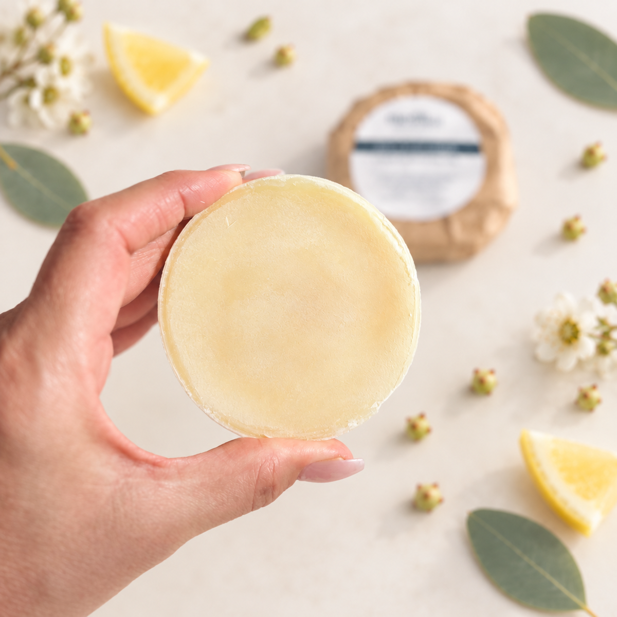 A hand holds a round, pale solid dish soap product, with a wrapped round product and lemon slices with small white flowers scattered in the background on a light beige surface. Made by Medika Organics 