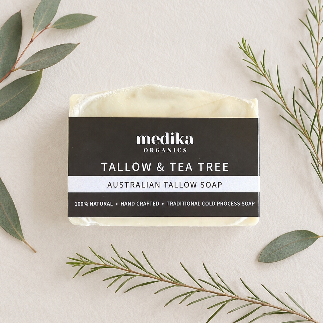 A bar of Medika Organics Tallow & Tea Tree Australian Tallow Soap is displayed in a flat-lay arrangement on a textured off-white background, surrounded by green eucalyptus and tea tree leaves.