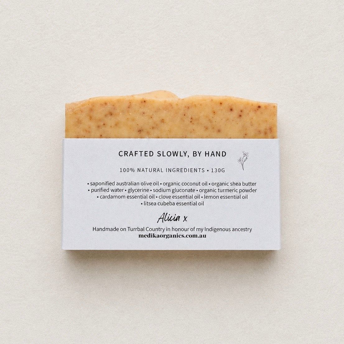 A single bar of handmade soap with a white paper label, photographed against a textured off-white background. The soap has a warm beige base with small dark speckles, and the label reads 'CRAFTED SLOWLY, BY HAND' and '100% NATURAL INGREDIENTS'.