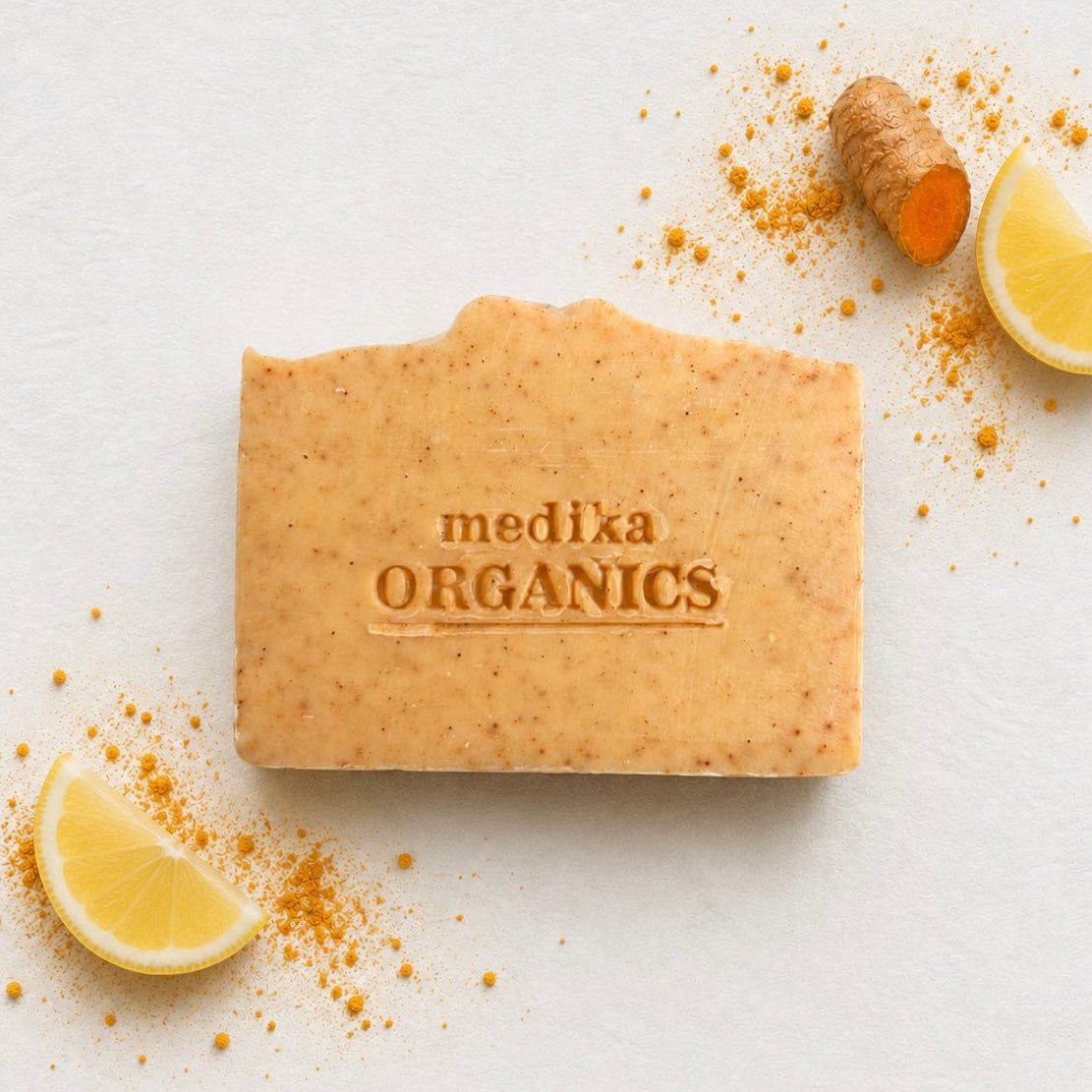 A rectangular bar of Medika Organics soap in a warm tan-orange color with visible speckles, placed on a light textured background with lemon slices, turmeric root, and loose turmeric powder around it. The soap is embossed with the brand name 'medika ORGANICS'.