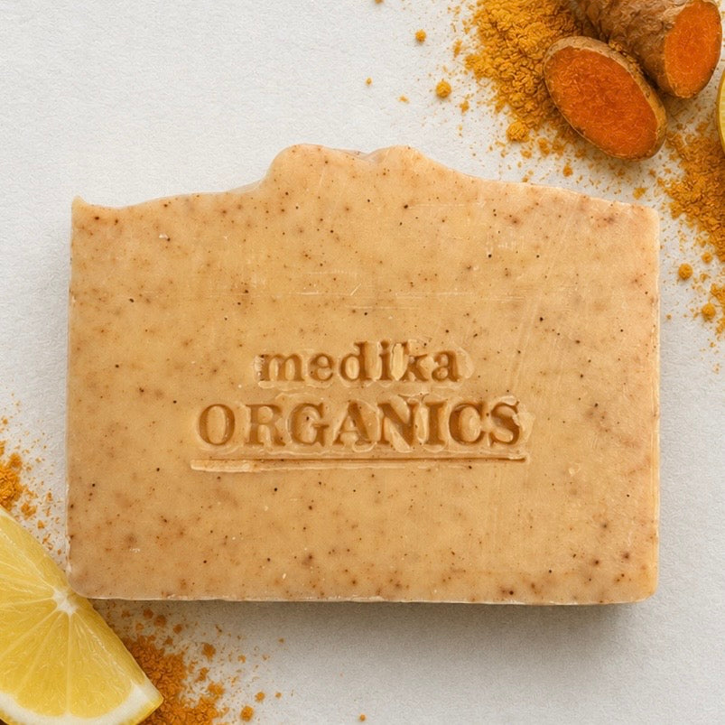 A rectangular bar of Medika Organics handmade soap with visible speckles, embossed with the brand name, styled on a white surface with turmeric root, turmeric powder, and a lemon slice.