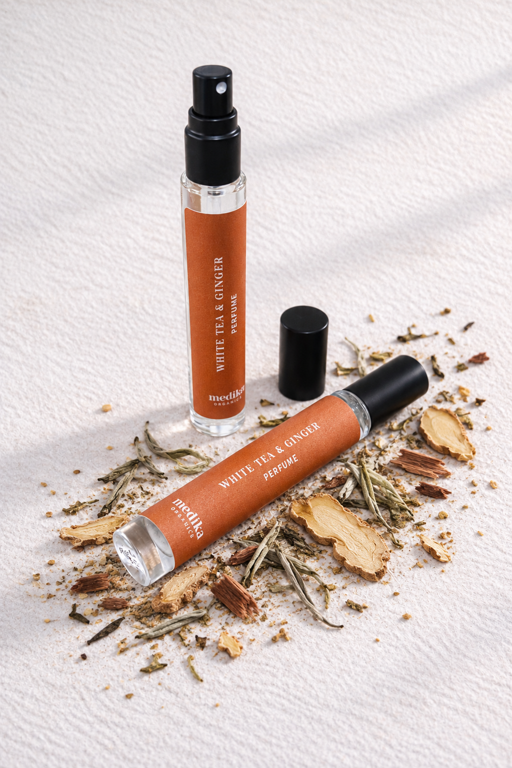 A clear cylindrical perfume bottle and matching roll-on bottle with orange labels reading White Tea & Ginger Perfume, arranged on a light textured surface with dried tea leaves and ginger slices around them.