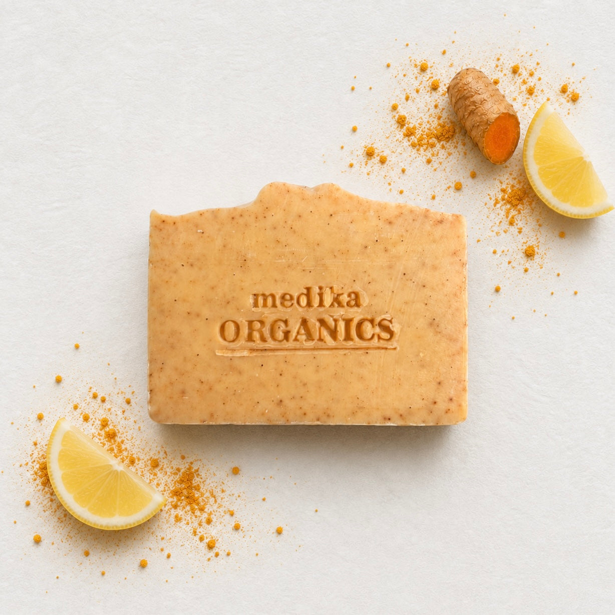 A rectangular bar of Medika Organics soap in a light tan color with visible speckles, placed on a white textured background with lemon slices, turmeric root, and turmeric powder scattered around it.