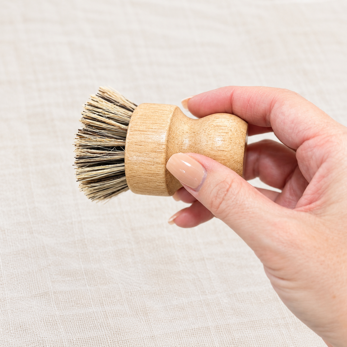 A hand holding a small wooden-handled cleaning brush with natural beige and dark brown bristles against a light beige textured fabric background.