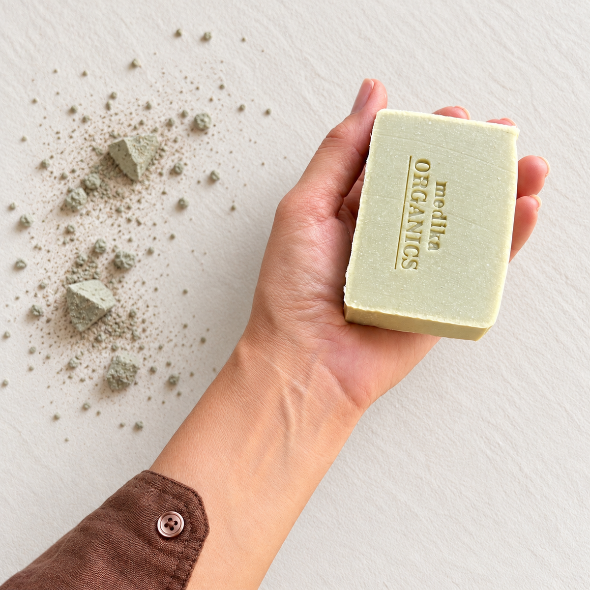 A hand holds a pale yellow-green rectangular bar of soap embossed with 'MEDIKA ORGANICS' against a light textured background with scattered marine clay.