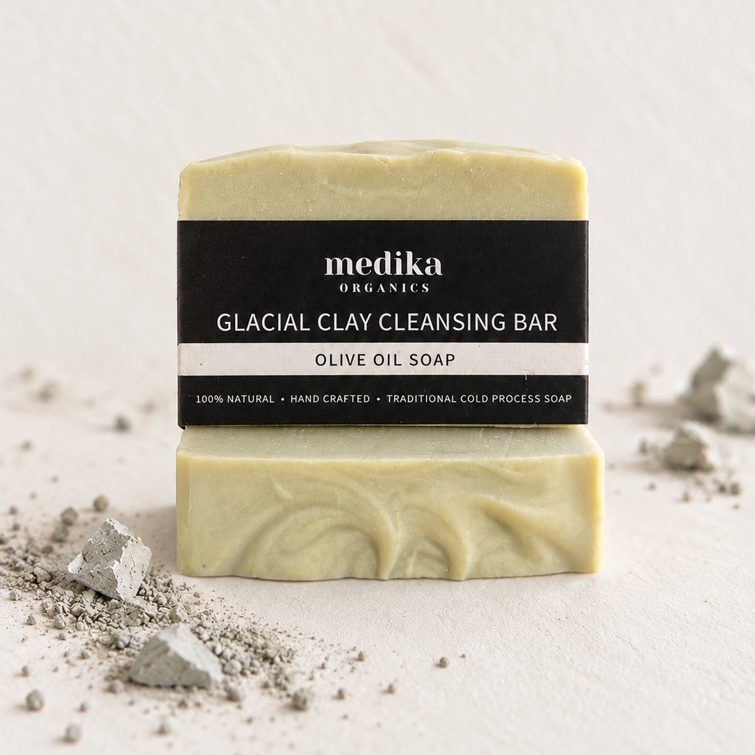A bar of Medika Organics Glacial Clay Cleansing Bar in Olive Oil Soap, featuring a black label with white text, set against a textured off-white background with scattered clay particles.
