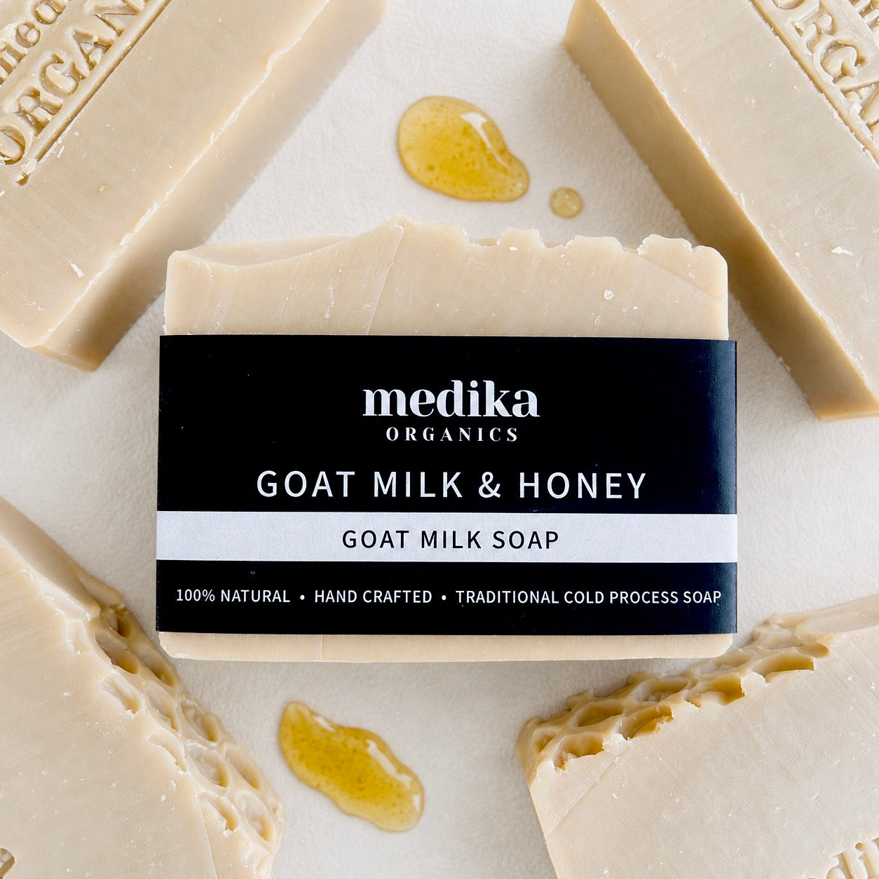 A bar of Medika Organics Goat Milk & Honey soap is centered on a light surface, surrounded by other soap bars and small pools of honey. The central bar has a black label with white text.