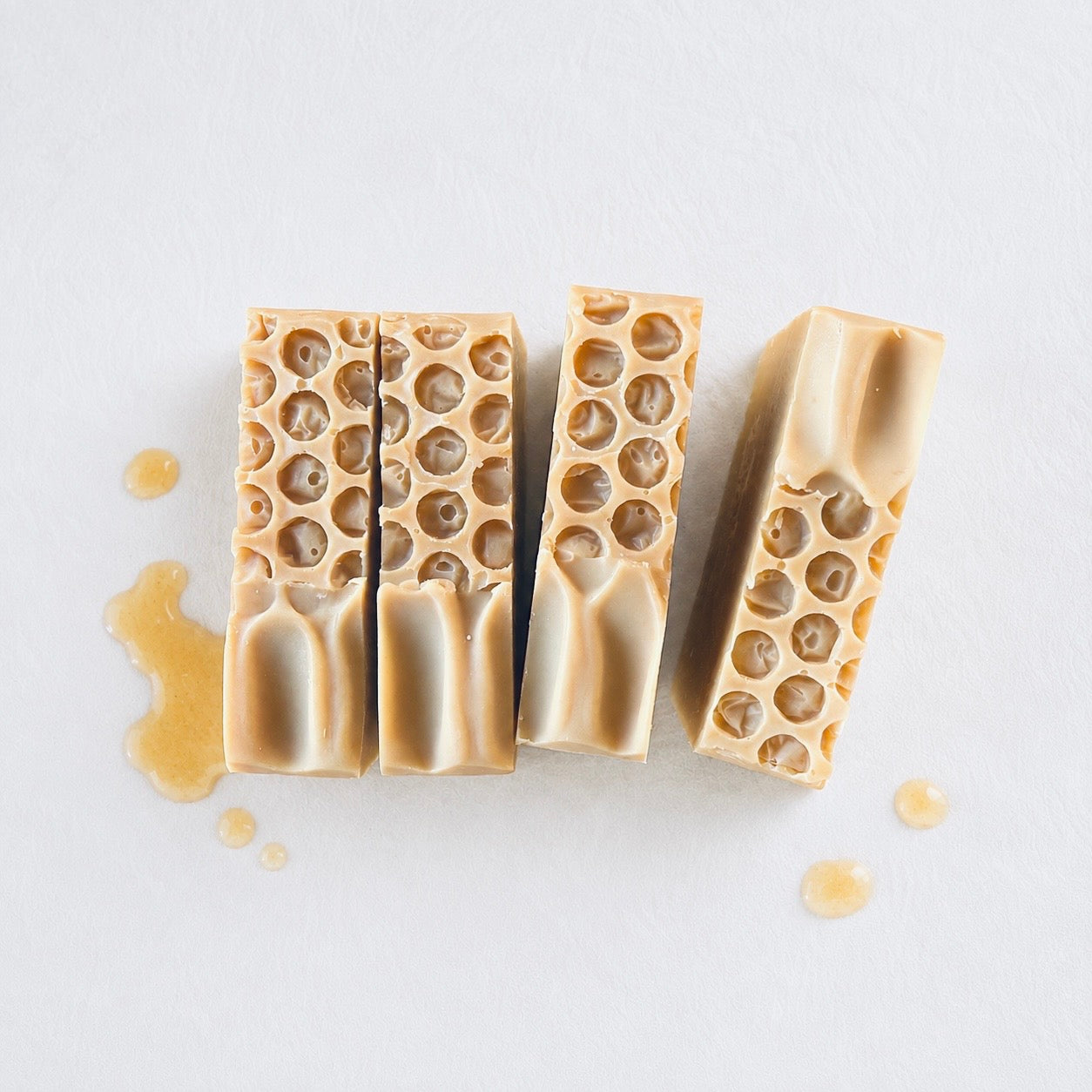 Four rectangular bars of honey and goat milk on a light gray background with a few drops of golden honey scattered around them.