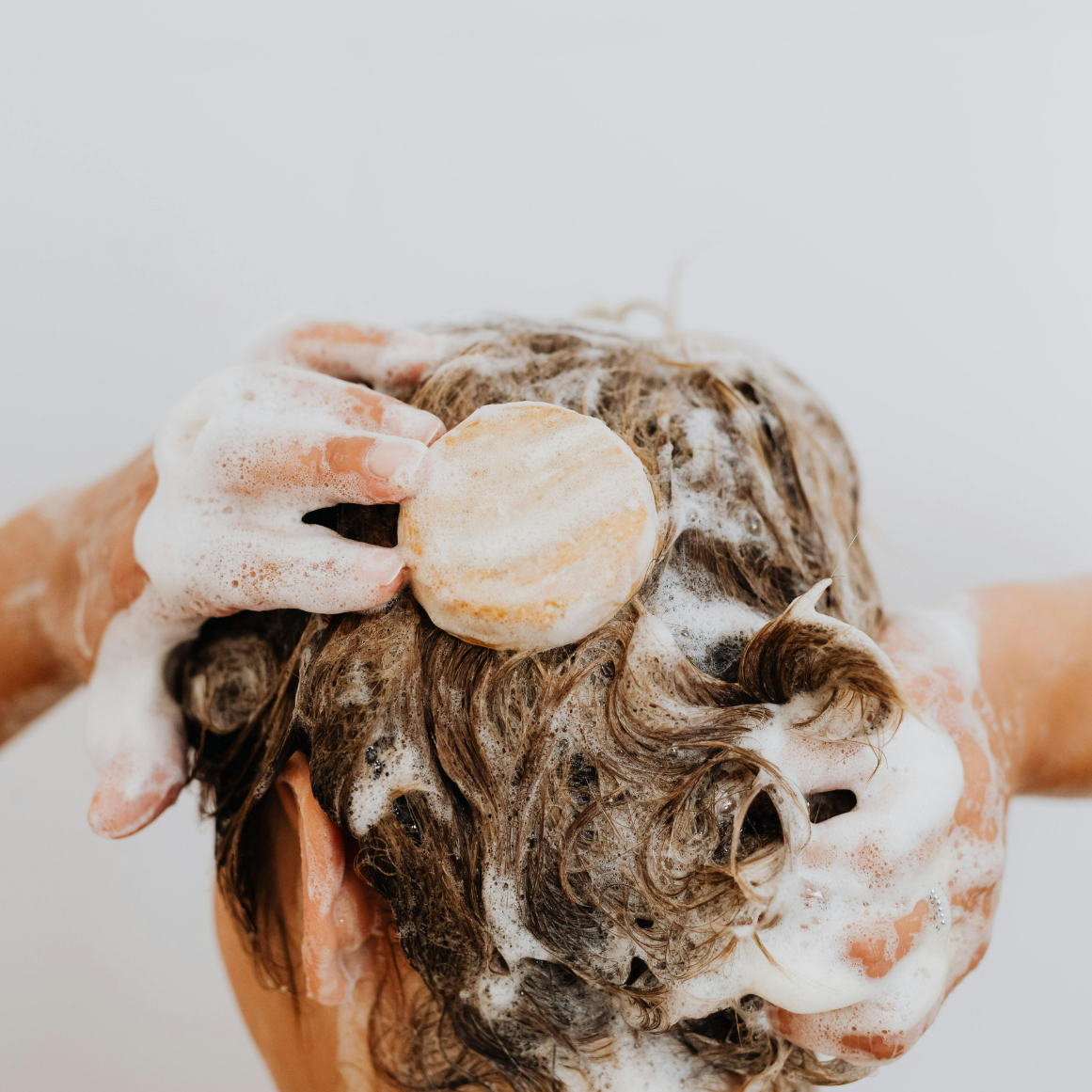 Person washing hair with a bar of shampoo against a neutral background