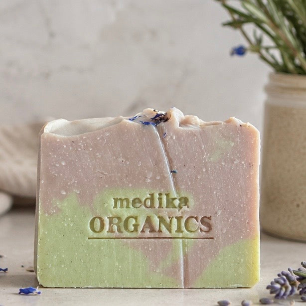 A handmade bar of soap from medika ORGANICS, featuring a marbled pink and light green color story with dried lavender buds on top, set against a rustic neutral background with a vase of greenery.