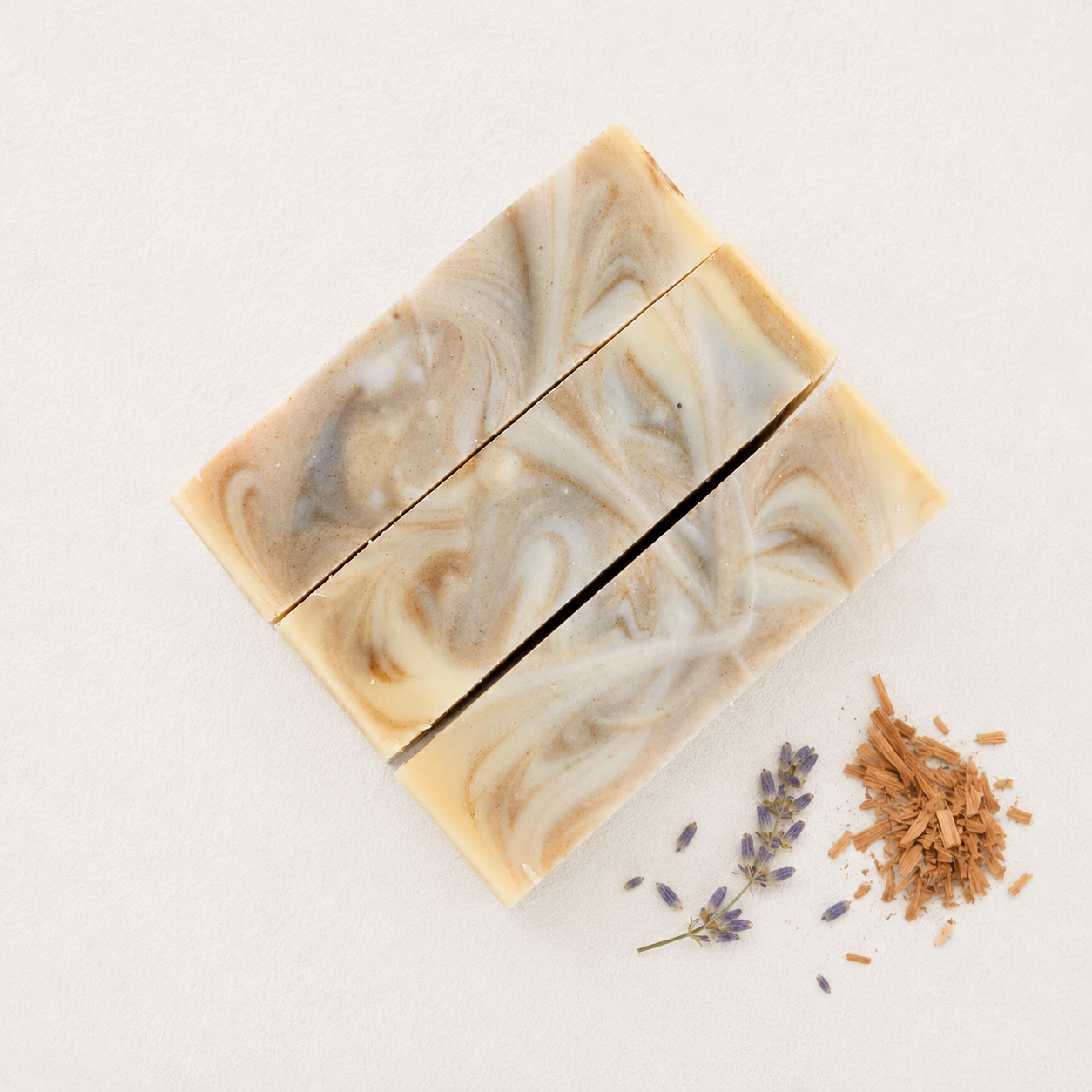 Three rectangular bars of marbled artisan soap with cream, beige, and gray swirls, arranged on a light neutral background with dried lavender and cedarwood botanicals beside them.