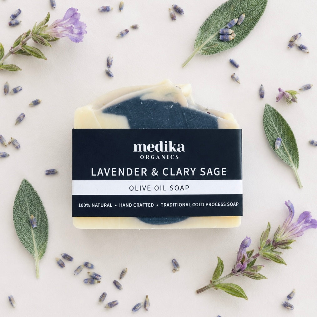 A bar of Medika Organics Lavender & Clary Sage Olive Oil Soap is centered on a light background, surrounded by fresh lavender sprigs, sage leaves, and scattered lavender buds. The soap has a marbled cream and dark teal-blue color, with a black paper label wrapped around it.