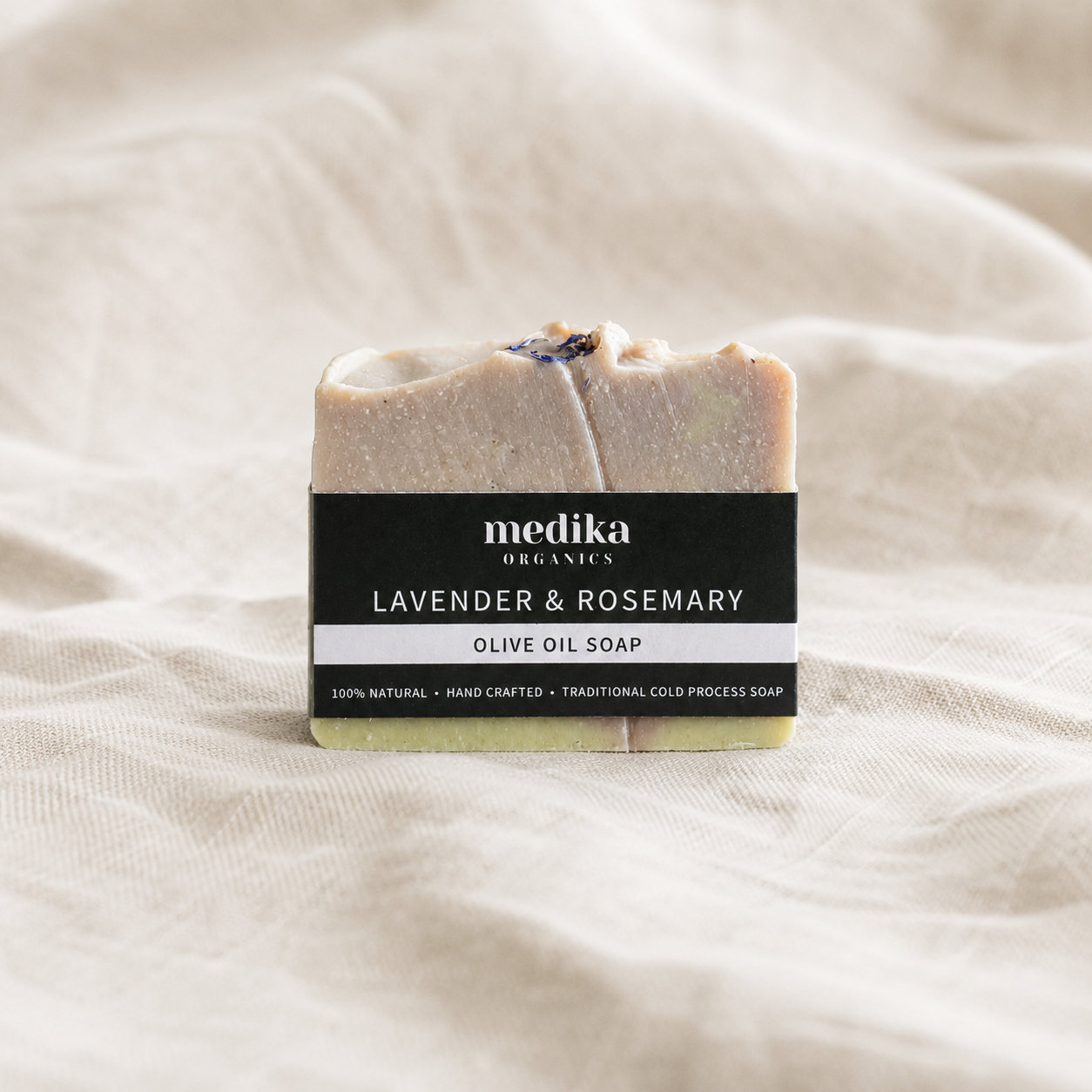 A single bar of Medika Organics Lavender & Rosemary Olive Oil Soap is centered on a soft, draped beige fabric background. The soap bar is a light, warm beige with a dark purple rosemary sprig embedded on top, wrapped in a black paper label with white text.