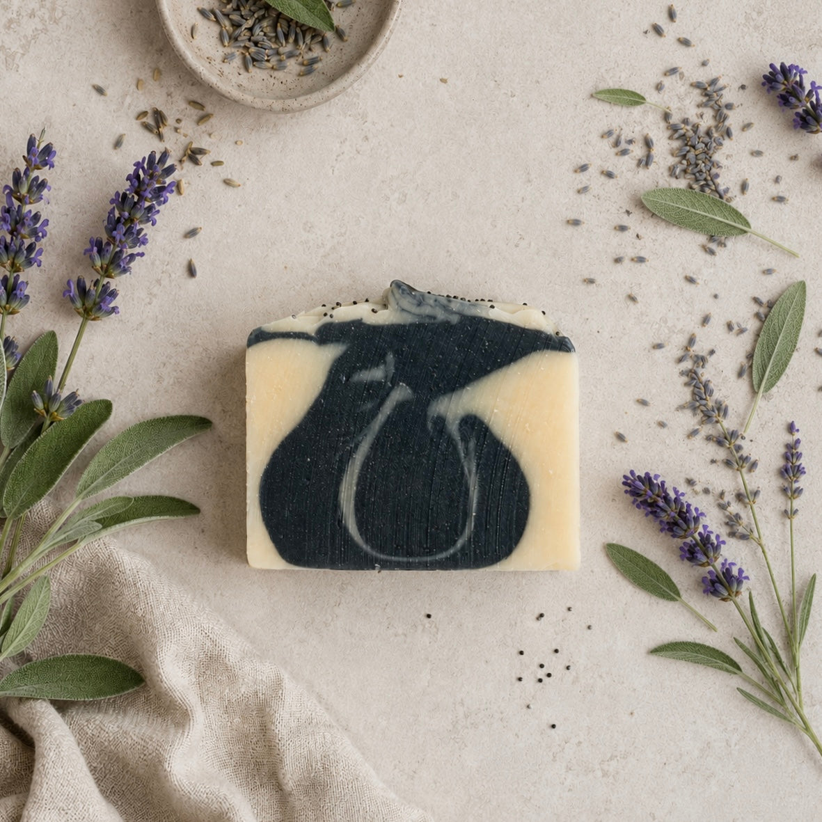 A rectangular artisan soap bar with a dark navy blue and cream marbled swirl pattern, styled on a textured beige surface with lavender sprigs, sage leaves, and a small bowl of dried lavender.