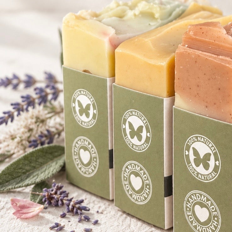Three handmade soap bars in olive green paper sleeves with butterfly logos, styled with lavender sprigs and a pink flower on a light textured surface.