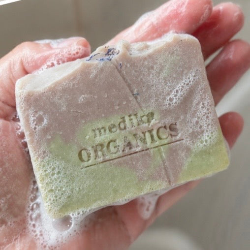 A hand holding a lathered bar of soap with visible foam. The soap is light green and beige with the brand name 'medLP ORGANICS' embossed on the front.