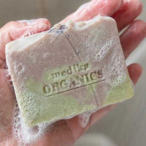 A hand holding a lathered bar of soap with visible foam. The soap is light green and beige with the brand name 'medLP ORGANICS' embossed on the front.