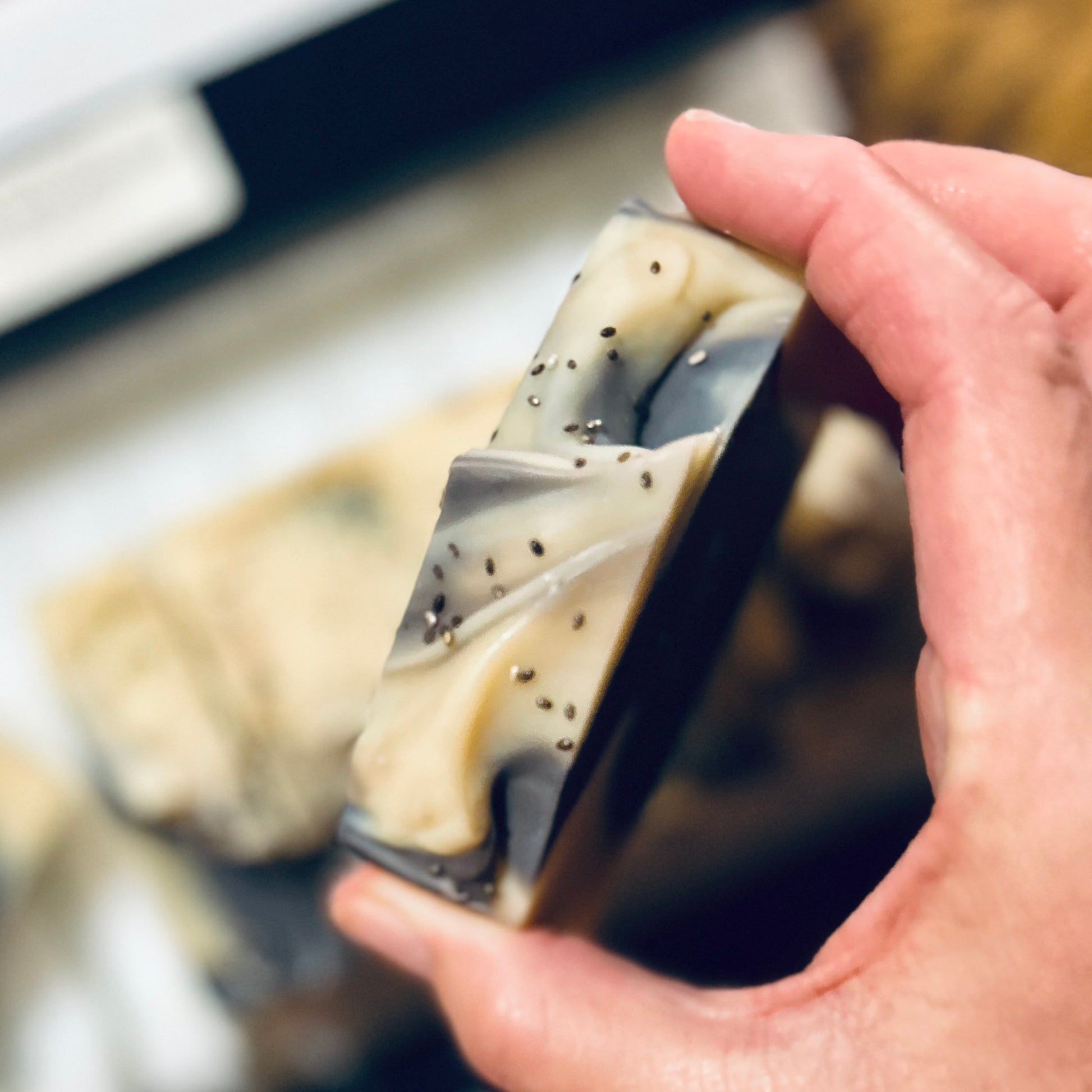 A hand holding a rectangular handmade soap bar with marbled beige, gray, and black swirls and small black seed-like inclusions, with more soap bars blurred in the background.