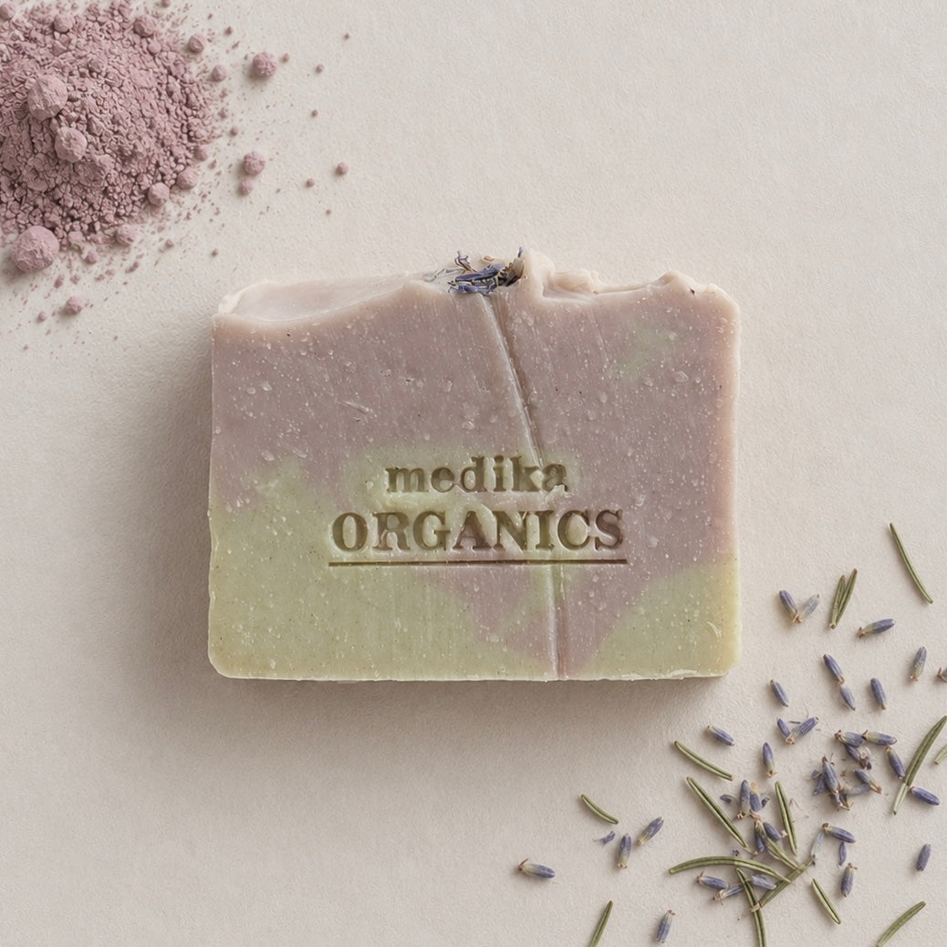 A rectangular bar of Medika Organics soap with a lavender and pale green color gradient, embossed with the brand name. The soap is styled on a neutral beige background with scattered dried lavender sprigs and a pile of lavender powder in the top-left corner.