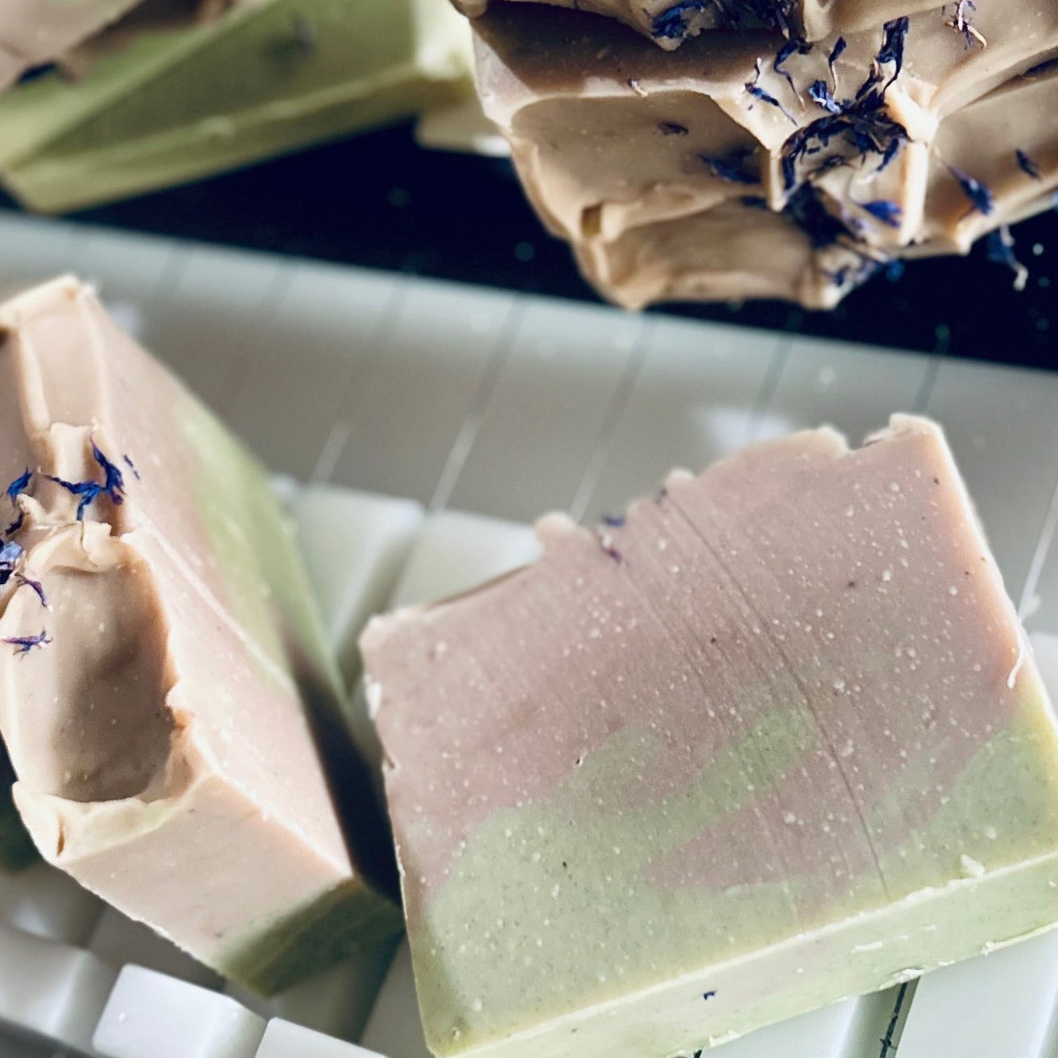Three handmade rectangular bars of soap resting on a white plastic soap mold. The soap features a marbled color transition from dusty pink to light green, with small dark blue dried flower petals embedded on the surface.