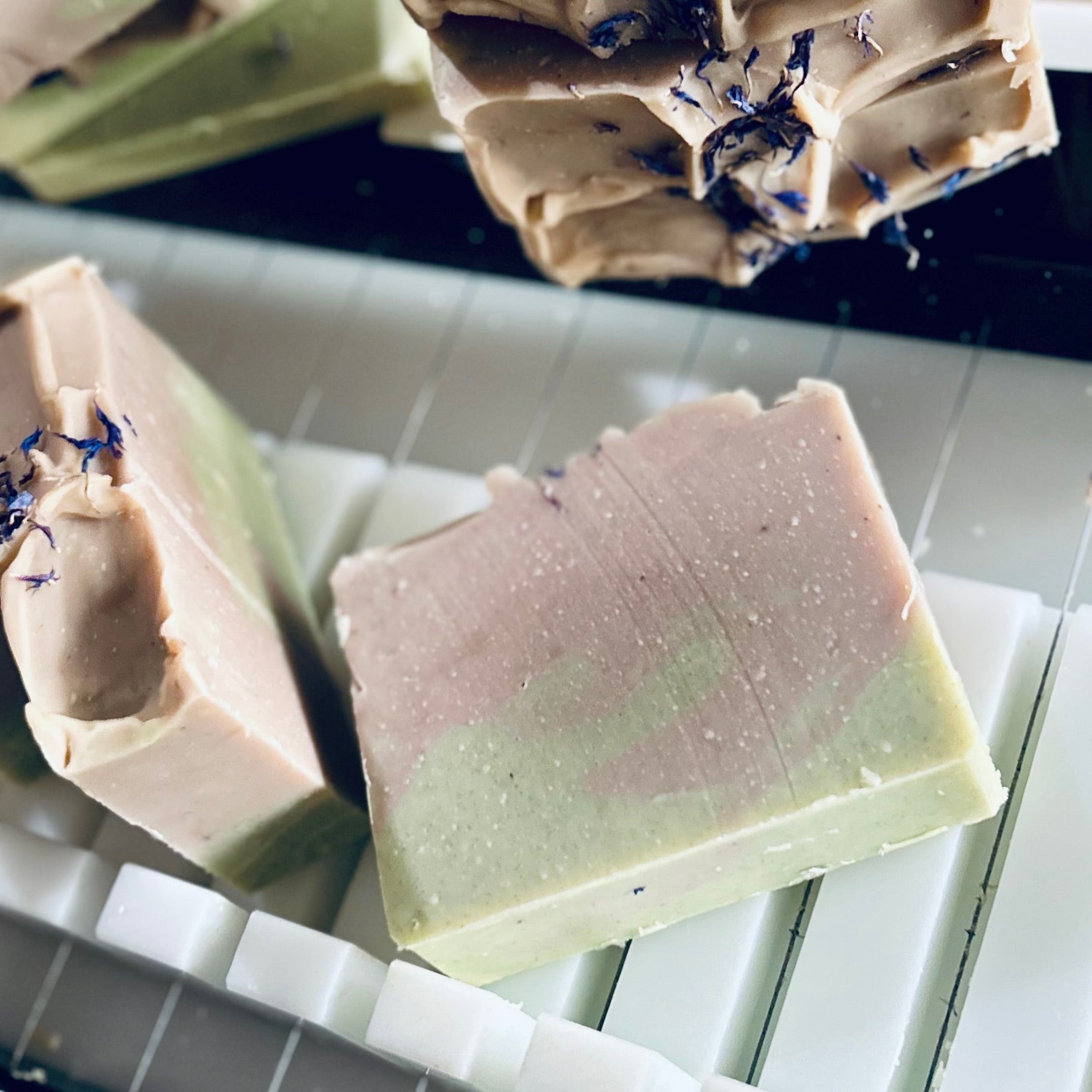 Three handmade rectangular bars of soap resting on a white plastic soap mold. The soap features a marbled color transition from dusty pink to light green, with small dark blue dried flower petals embedded on the surface.