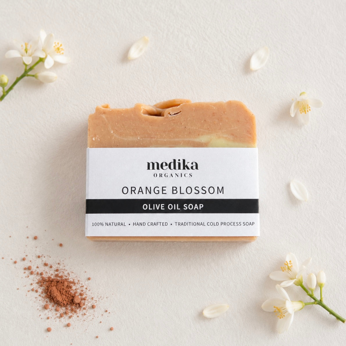 A bar of Medika Organics Orange Blossom Olive Oil Soap is displayed on a textured off-white background, styled with small white blossoms, scattered petals.