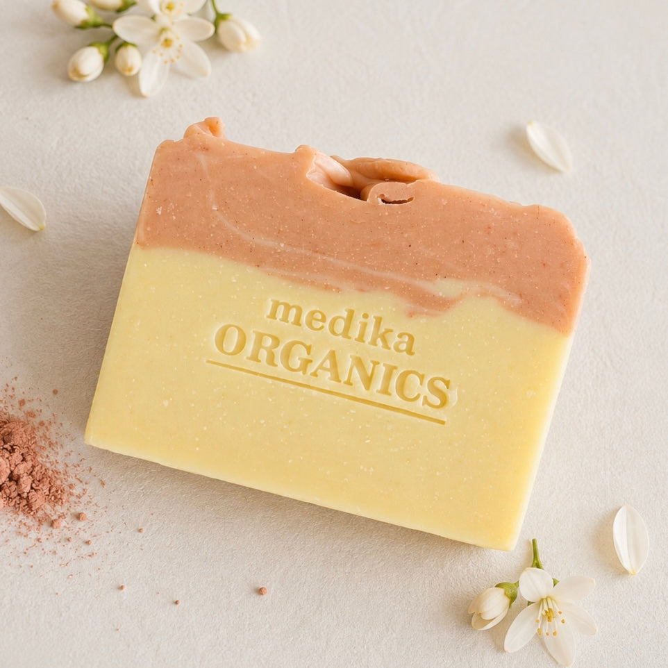 A rectangular bar of handmade soap from Medika Organics, featuring a two-tone design with cream lower half and a peachy-pink upper half. The soap is embossed with the brand name and is styled on a textured light beige surface with scattered white blossoms and a touch of pink clay powder.