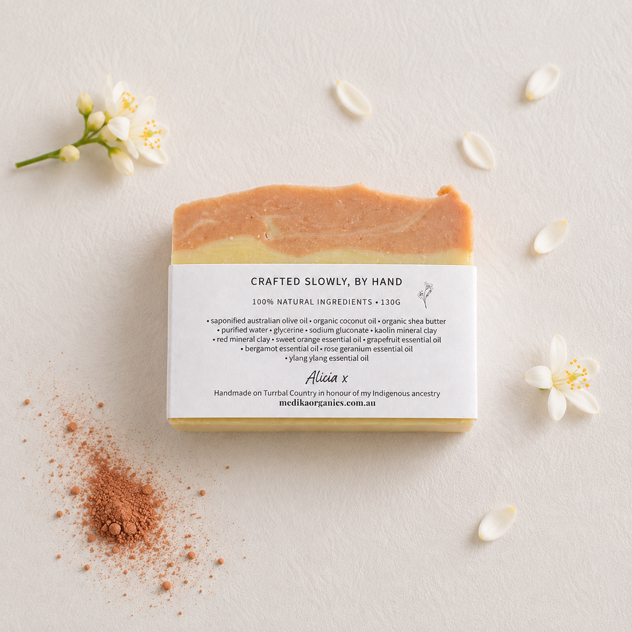 A rectangular bar of Medika Organics handmade soap with a white paper label, styled on a light beige textured background with scattered white flower petals.