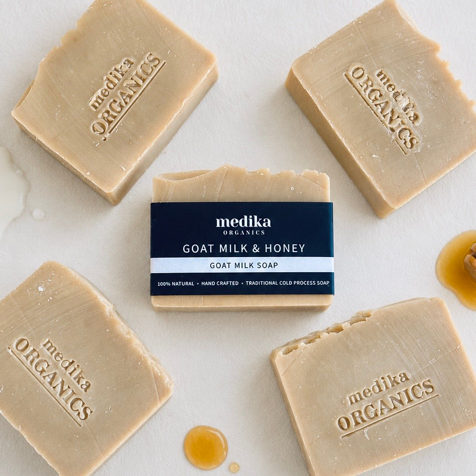 Four bars of Medika Organics Goat Milk & Honey soap arranged on a light marble surface, with one bar centered and its dark navy label facing forward. Honey is lightly drizzled around the bars.