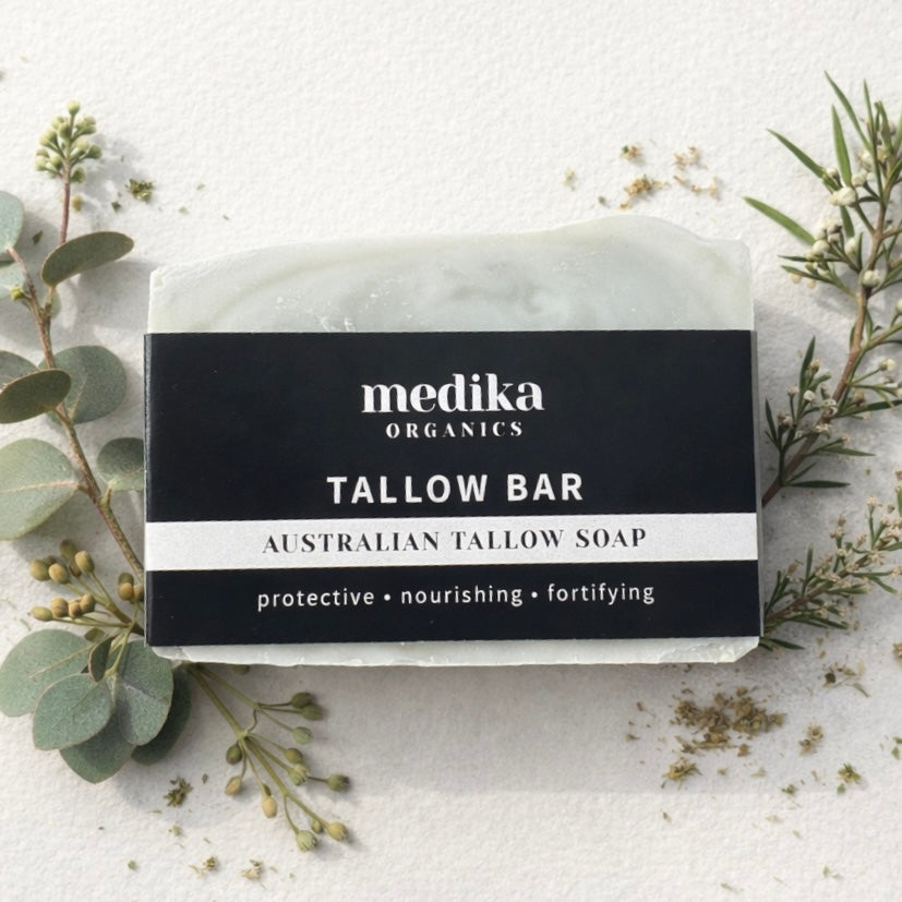 A bar of Medika Organics Australian Tallow Soap wrapped in a black and white paper band, styled on a textured white surface with botanicals. 